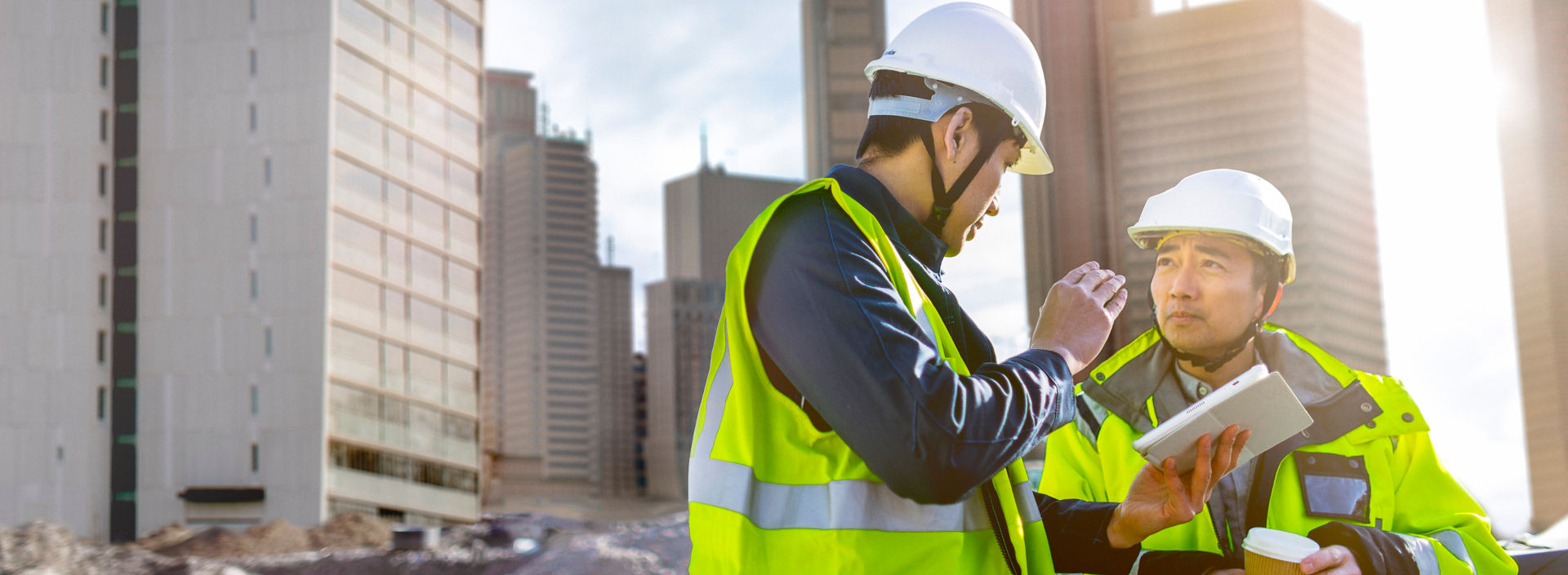 Two men in hi-vis jackets have a conversation on a building site while looking at a tablet.
